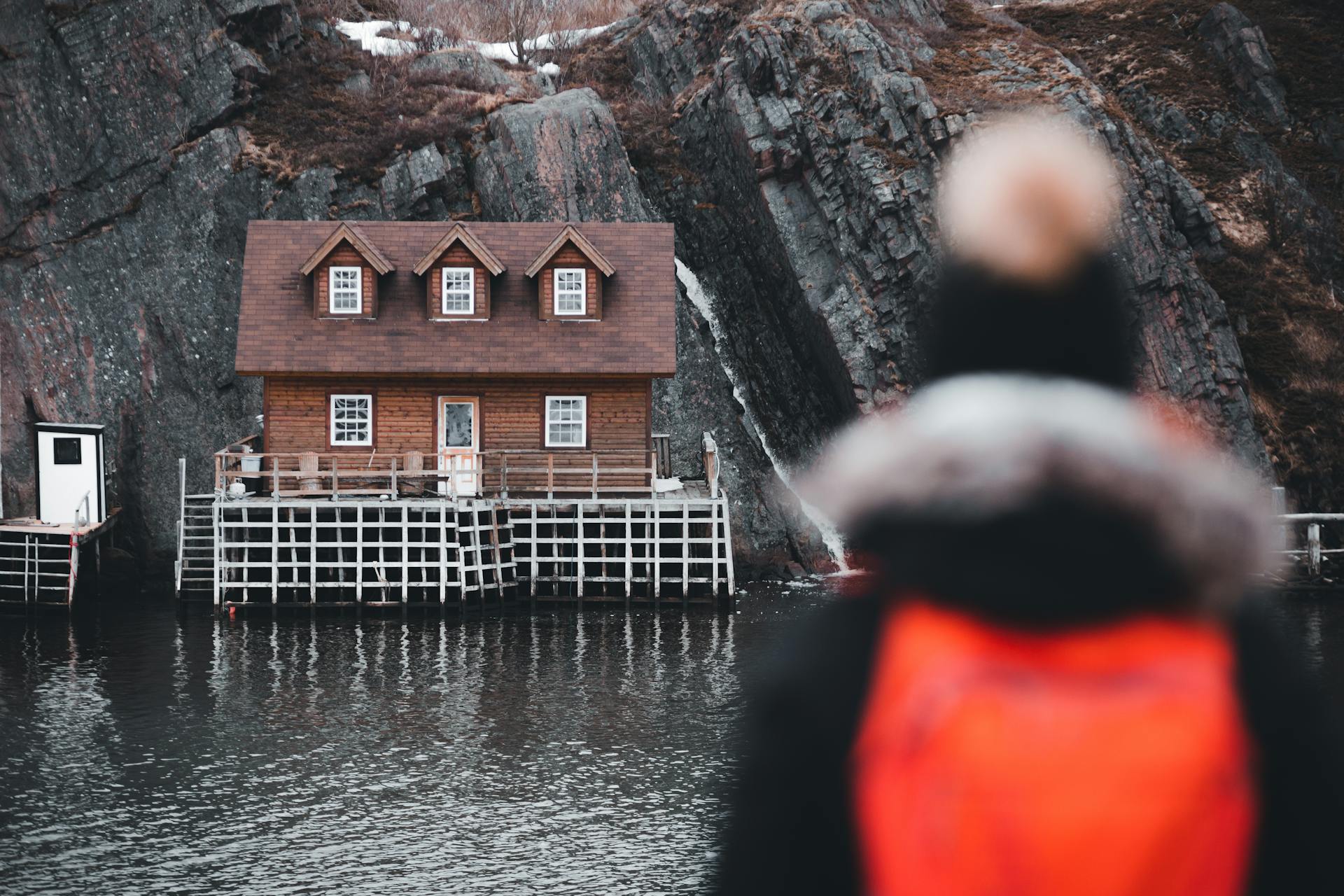 A serene winter scene featuring a cozy lakehouse by rocky cliffs and a person with a red backpack.