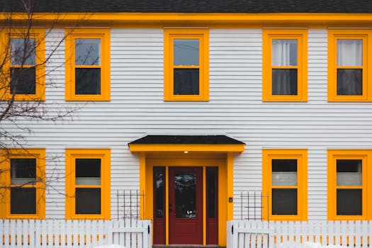 A vibrant yellow-trimmed house with a red door, fence, and multiple windows.