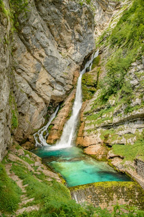 Powerful waterfall streaming through rough rocky cliff and flowing into calm lake with turquoise water on sunny day