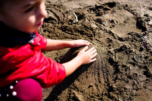 From above of crop little girl in colorful clothes squatting and playing with wet sand on beach on sunny day