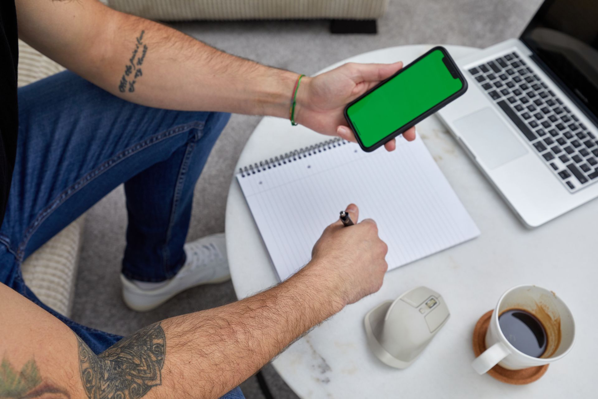 Man taking notes with phone, laptop, and coffee in a casual workspace