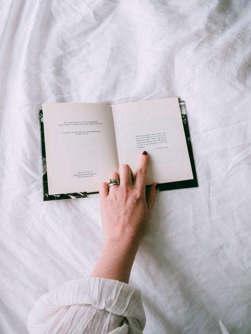 Free A hand with manicured nails pointing at text in an open book on a white bedspread. Stock Photo