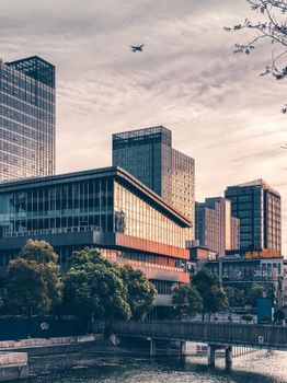 Airplane flying over modern buildings with glass mirrored walls located on river shore against cloudy sky during sunset