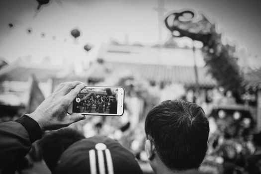 A monochrome image of a parade being recorded on a smartphone during a festival.