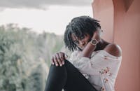 一位年轻、沉思的非裔美国女性侧身倚在阳台上，背后是模糊的热带树木，她留着非洲发型，穿着休闲服，正看向镜头