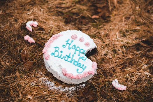 A partially eaten birthday cake lies on the ground outdoors, surrounded by pine needles.