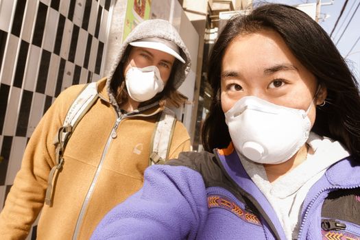 Two women wearing face masks, walking outdoors during daylight, reflecting pandemic safety measures.