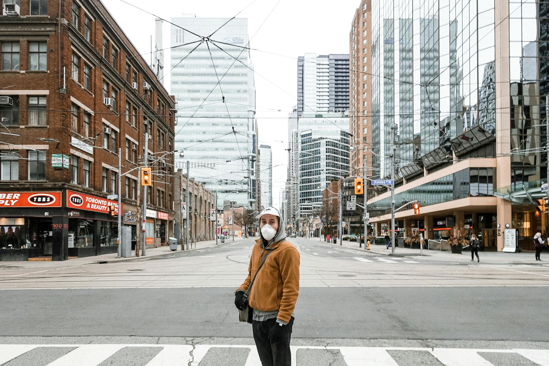 A person wearing a mask stands on a Toronto street surrounded by modern and historic buildings.