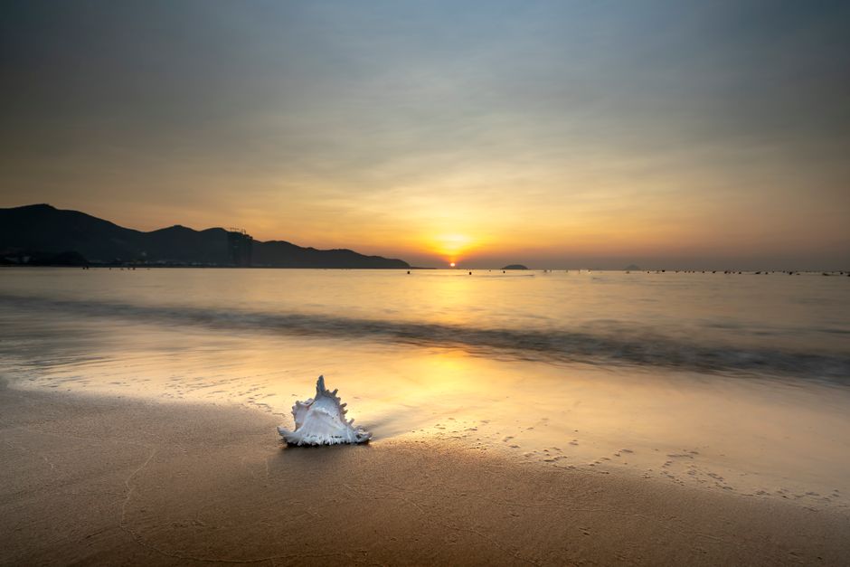 A serene sunset view over the ocean with a seashell on the sandy beach, framed by distant mountains.
