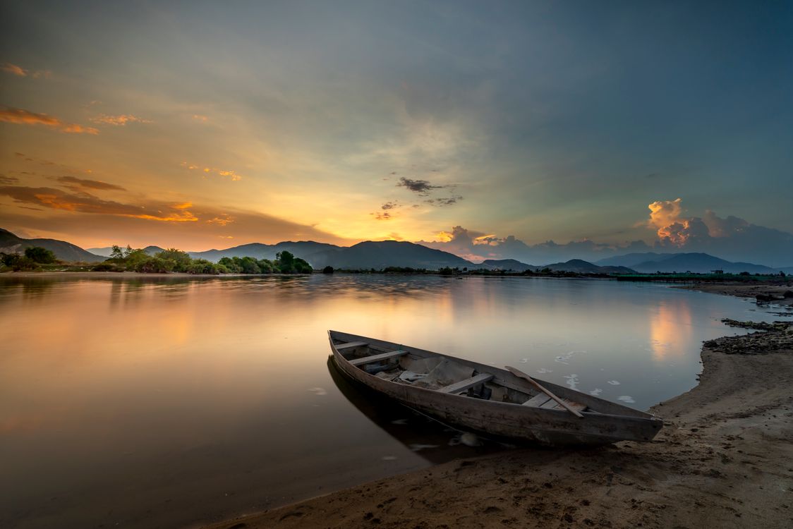 Free A beautiful sunset view over a tranquil lake with a solitary wooden rowboat on the shore. Stock Photo