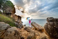 日落时分，一名戴帽子的女子撑着彩虹伞，站在海边风景优美的岩石悬崖上