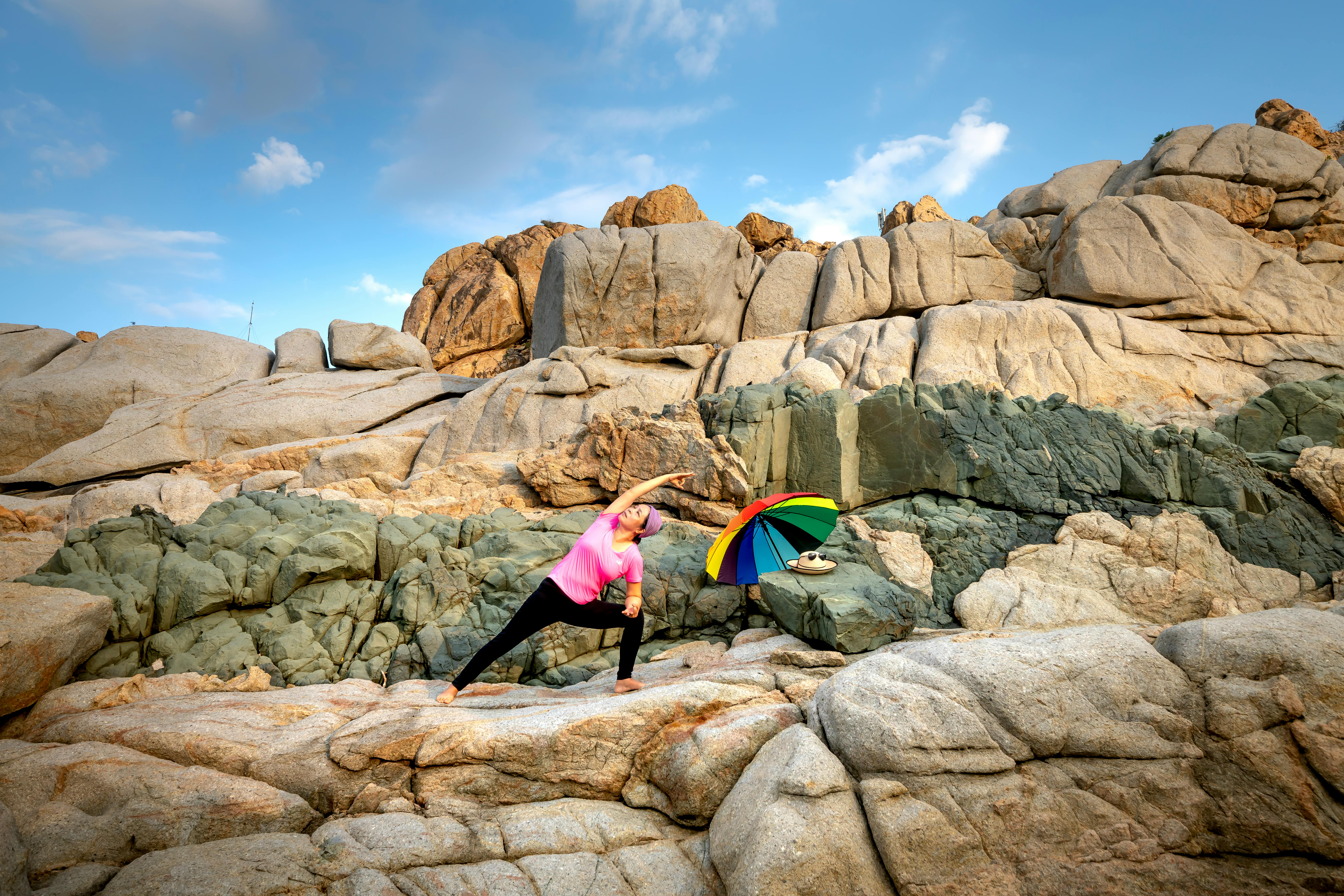 一名女子在晴朗的蓝天下，撑着彩色伞，在岩石地形上伸展身体做瑜伽