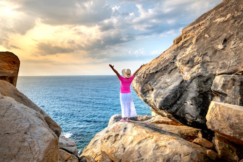 Free Woman in Pink Top and White Skirt Standing on Rock Formation Near Body of Water Stock Photo
