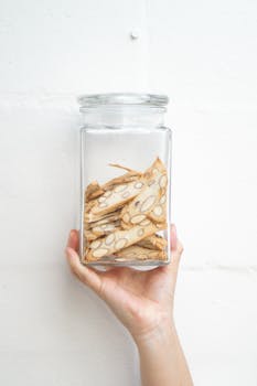 Hand holding a glass jar filled with almond biscotti, set against a white wall background.