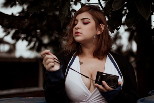 Trendy woman with makeup and tattoos posing outdoors with a camera.