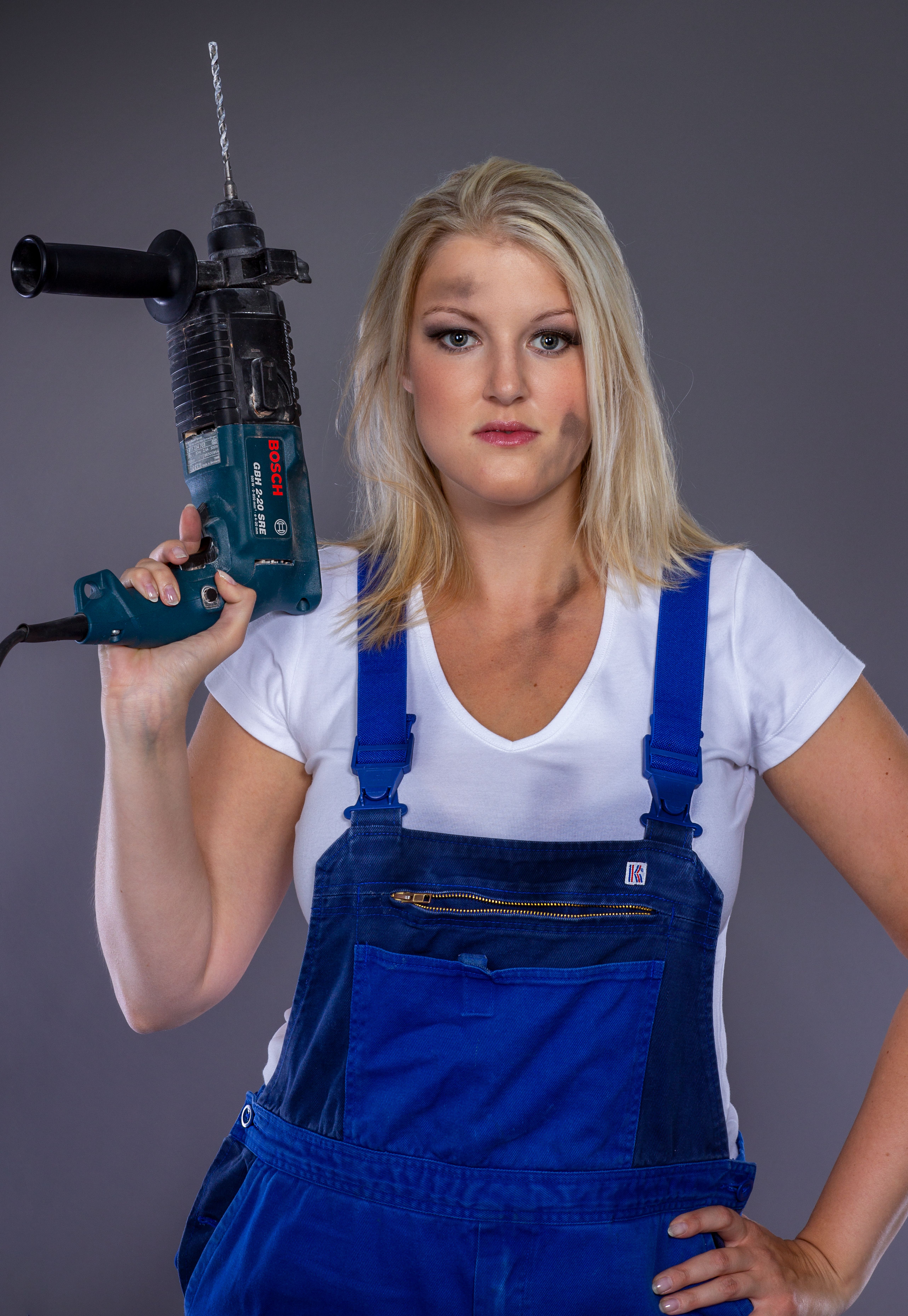 Portrait of a confident woman holding a power drill, wearing overalls and a white t-shirt in a studio setting.