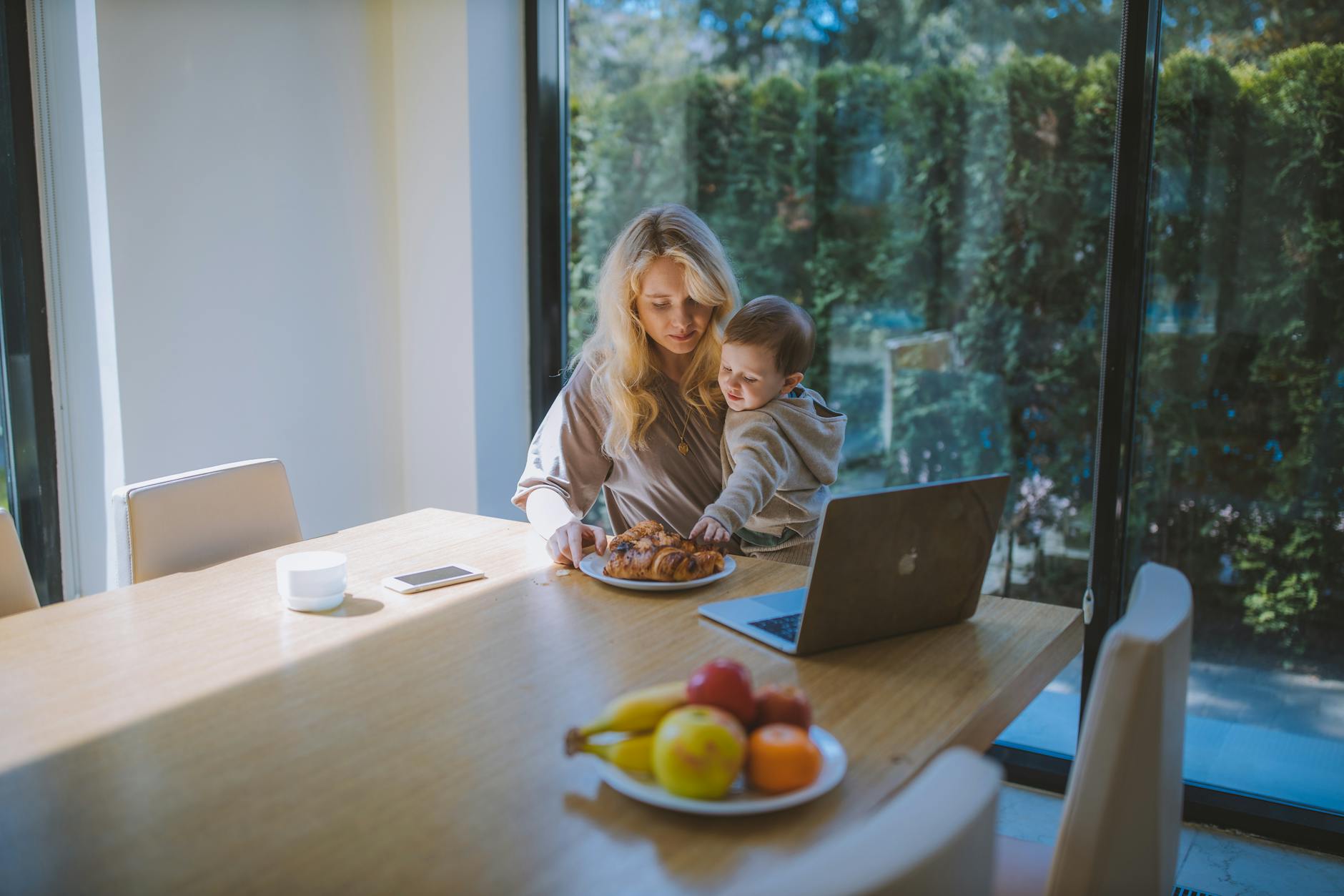 A mother shares breakfast with her child at a sunlit table, with a laptop and fruit bowl present.