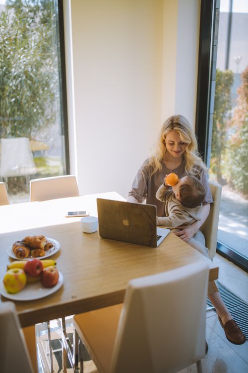 Free A blonde mother multitasks with her baby and laptop, enjoying breakfast in a bright home setting. Stock Photo