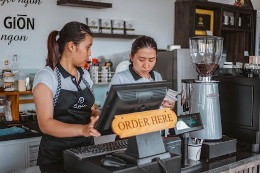 Two baristas work at a coffee shop counter with a cash register and equipment.