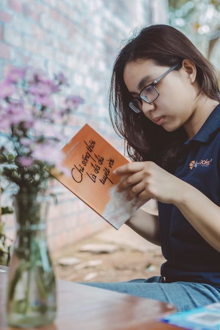 Young woman with glasses enjoying reading outdoors, surrounded by nature and flowers.