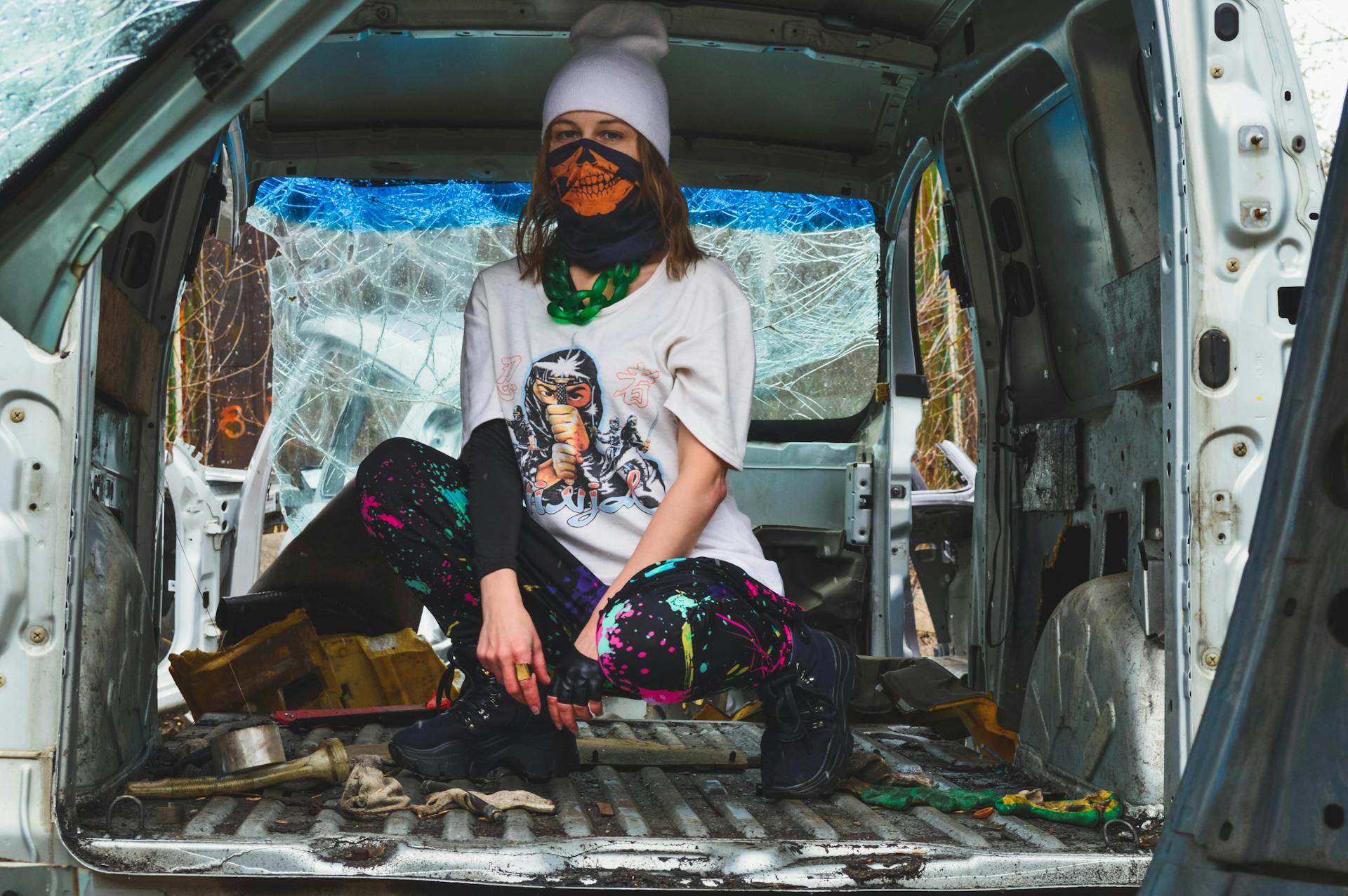 Full body of anonymous female teenager in informal clothes covering face with handkerchief and sitting on haunches in trunk of dirty broken car