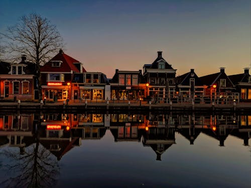 Traditional residential houses and leafless tree on shore of calm lake reflecting in water during sunset