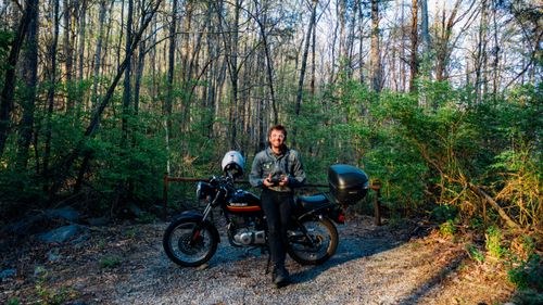 Free Man in forest with motorcycle, holding a remote, smiling at the camera. Stock Photo