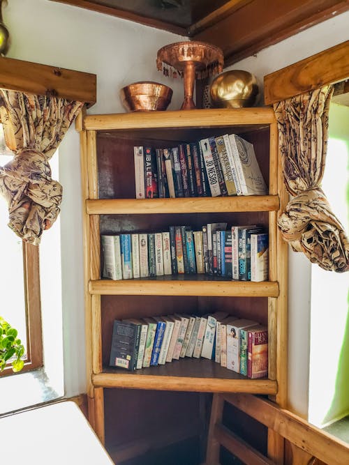 Vintage wooden shelf with various books and old  fashioned vases placed near windows in cozy house