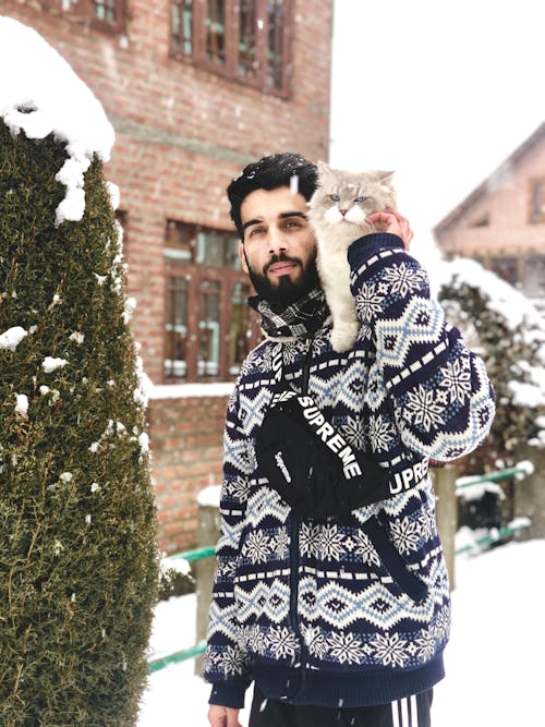 Young calm bearded Hispanic male in trendy warm clothes standing near brick building in village with cute fluffy cat on shoulder and looking at camera on snowy day