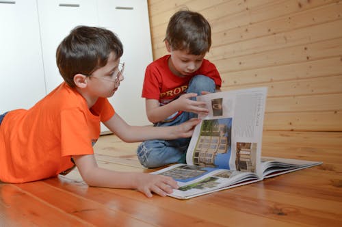 Free Curious smart little siblings in casual clothes sitting on wooden floor and reading interesting book at home Stock Photo