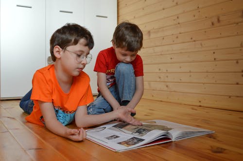 Free Cute little brothers in casual clothes reading interesting fairytale sitting on wooden floor while spending free time together at home Stock Photo