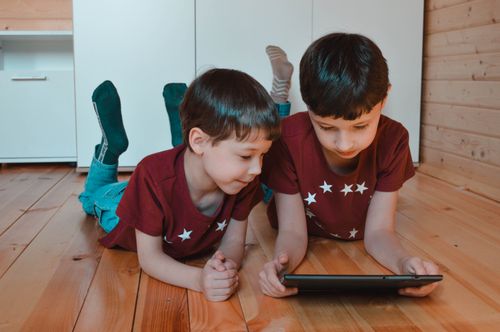 Free Adorable little siblings in similar clothes lying on wooden floor and watching video on tablet while spending weekend together at home Stock Photo