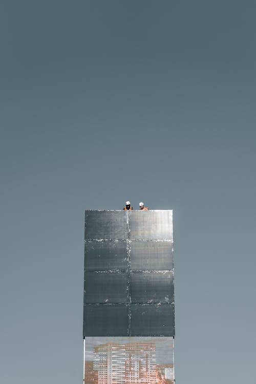 From below of anonymous distant engineers in helmets working on rooftop of modern skyscraper with glass mirrored walls against cloudless blue sky