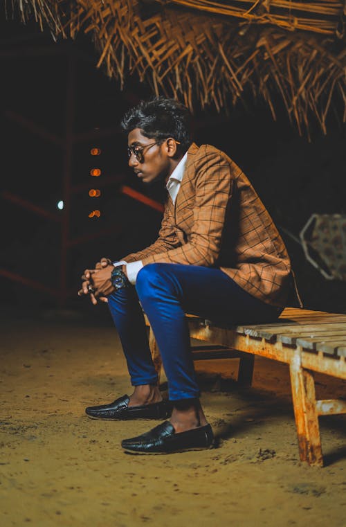 Full body side view of senior Indian man in trendy suit and sunglasses sitting under thatched roof