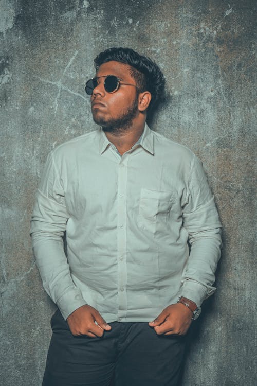 Confident ethnic male wearing modern sunglasses white shirt and black pants standing against gray wall and looking away