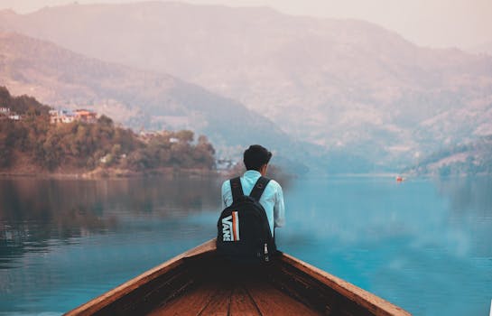 A man with a backpack sits on a boat, reflecting by a serene mountain lake.