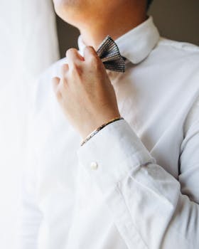 A man wearing a formal white shirt adjusts his bow tie indoors, illuminated by natural light.