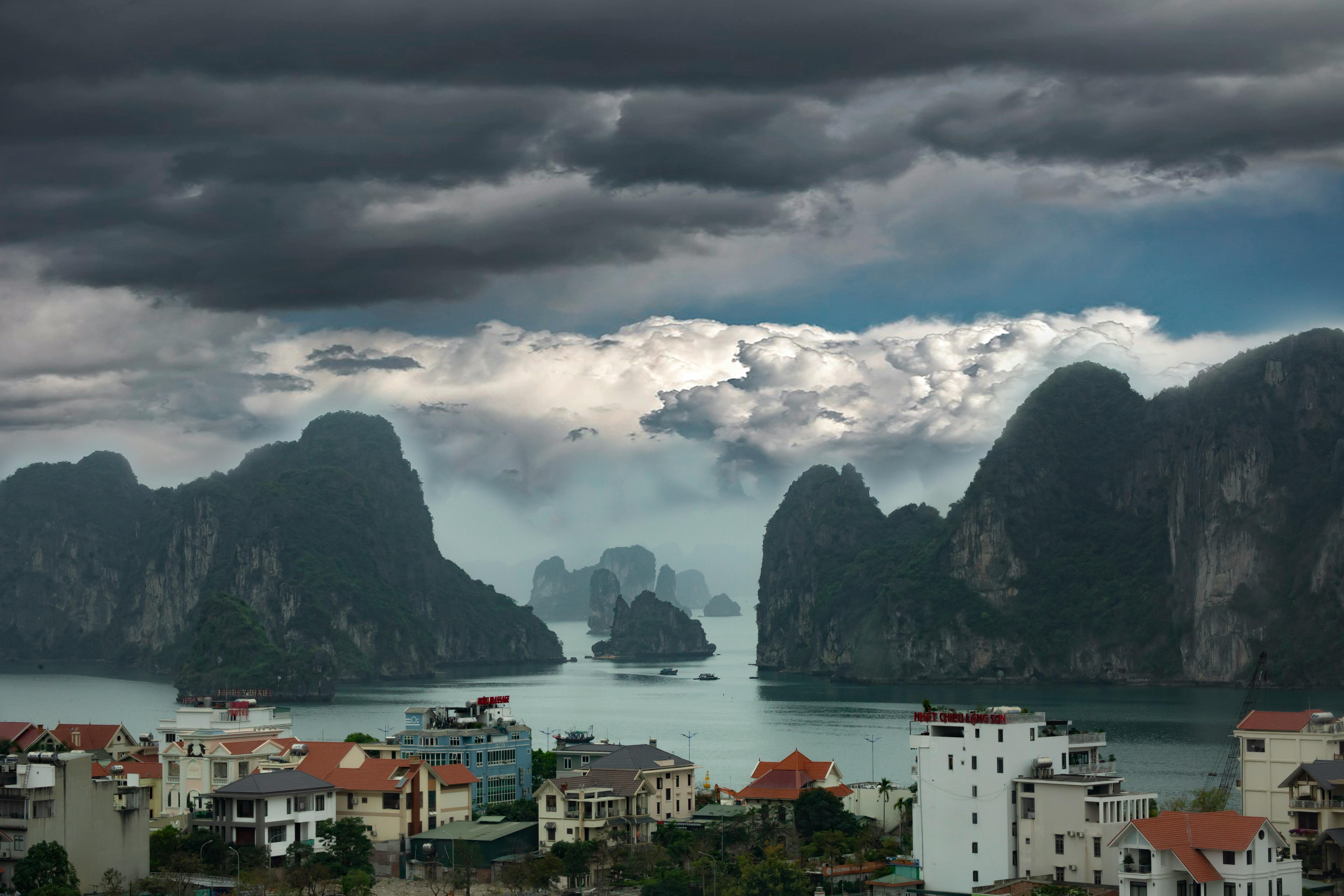 High angle of residential houses and of rocky cliffs covered with green plants in middle of sea under dark stormy sky