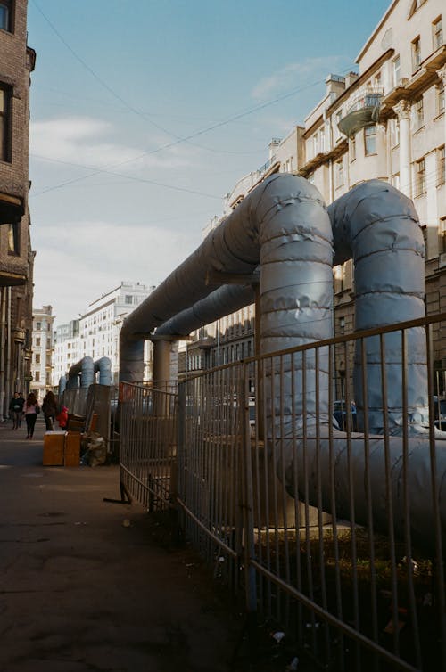 Water tubes on asphalt walkway near metal fence and modern multistage dwelling buildings under cloudy sky on city street in daylight