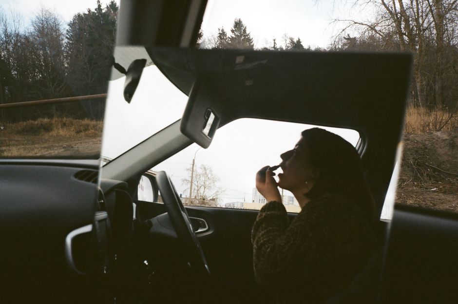 Through glass side view of anonymous female making up lips with lipstick while sitting with open mouth and looking in mirror in automobile near trees in countryside in autumn