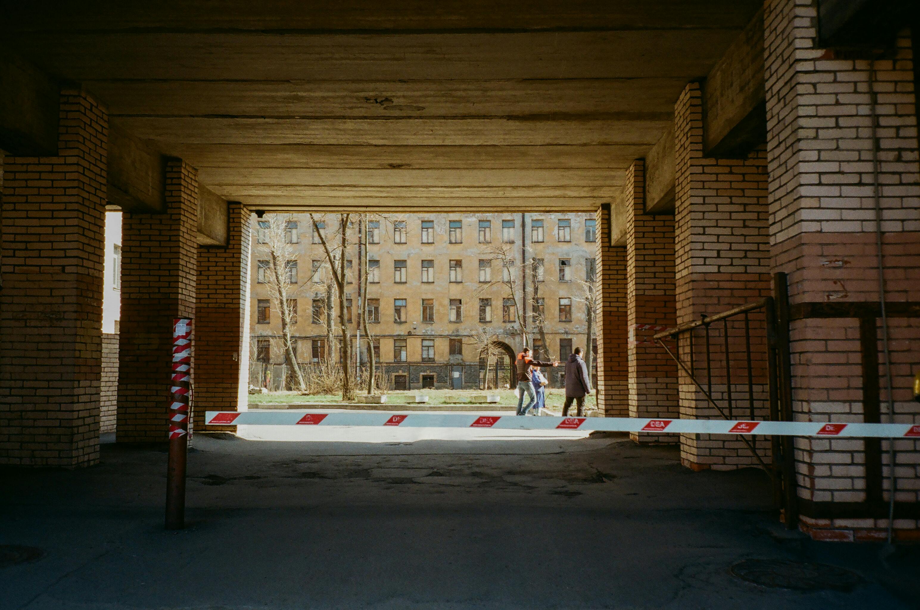 Asphalt pathway between contemporary buildings with brick wall near roadblock and dwelling multistage house in city with unrecognizable people walking on street
