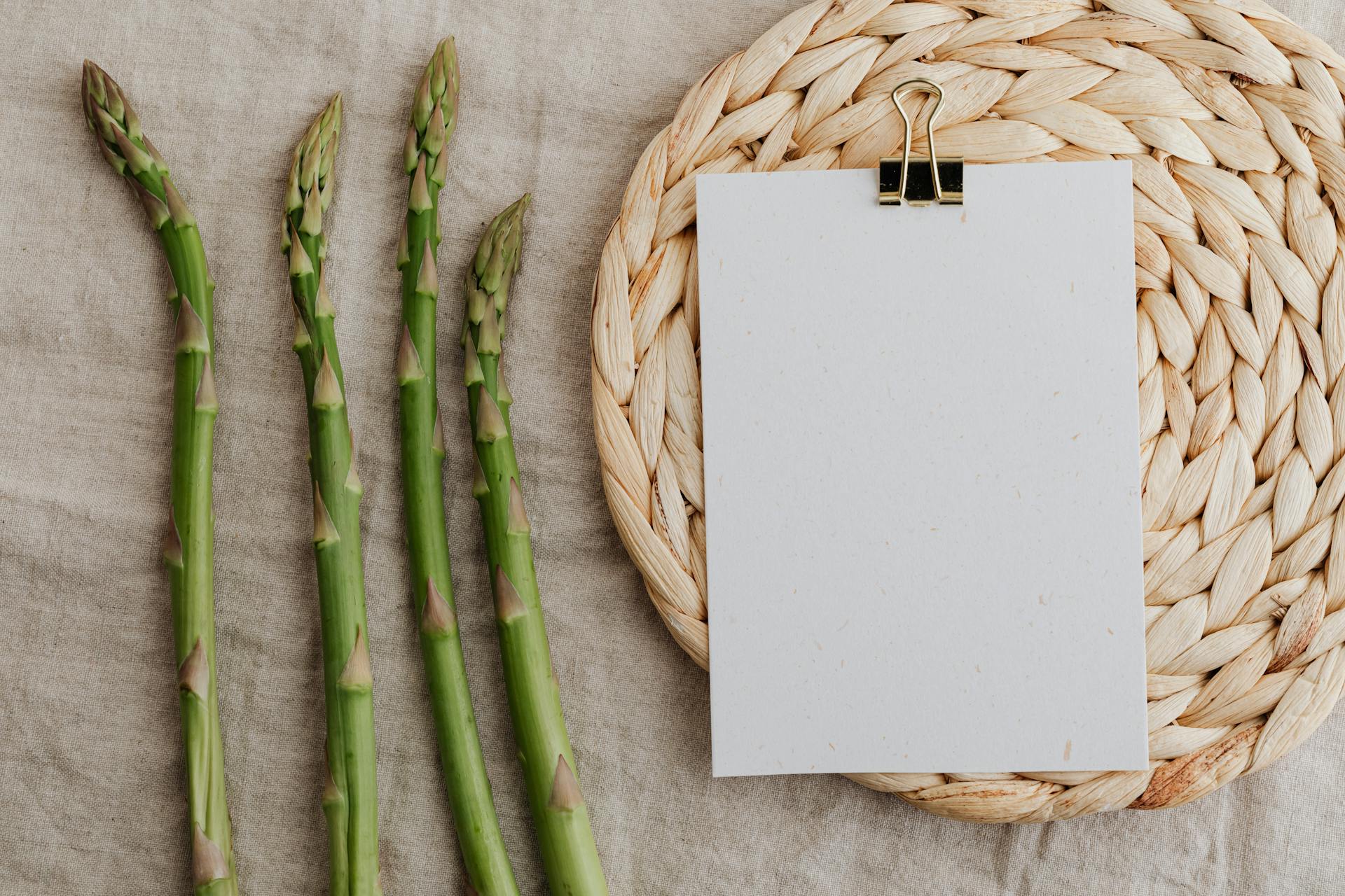 Fresh asparagus arranged on rustic fabric with a blank paper on a woven mat, perfect for culinary themes.