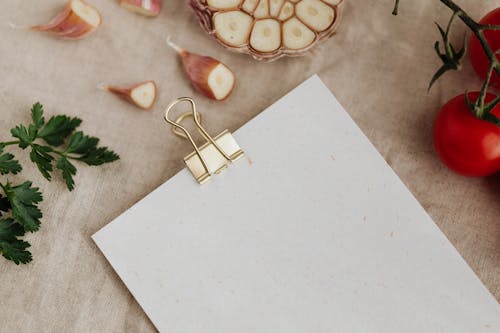 Top view of empty sheet of paper fixed with metal clip and placed near garlic gloves and ripe tomatoes with parsley on linen towel