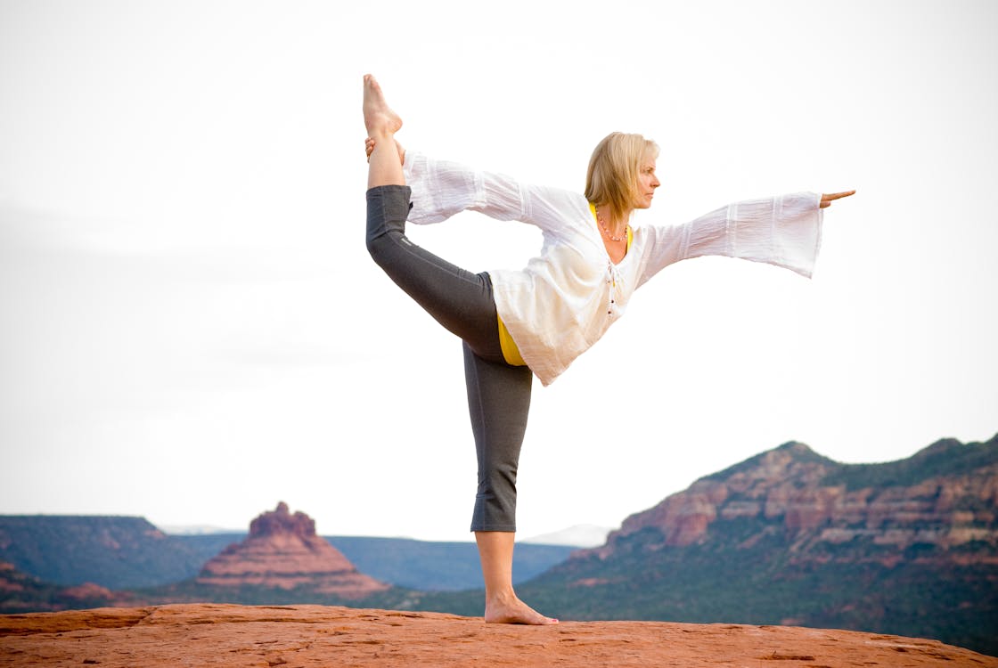 Free Woman in White Long Sleeve Shirt and Black Pants Standing on Brown Rock Formation Stock Photo