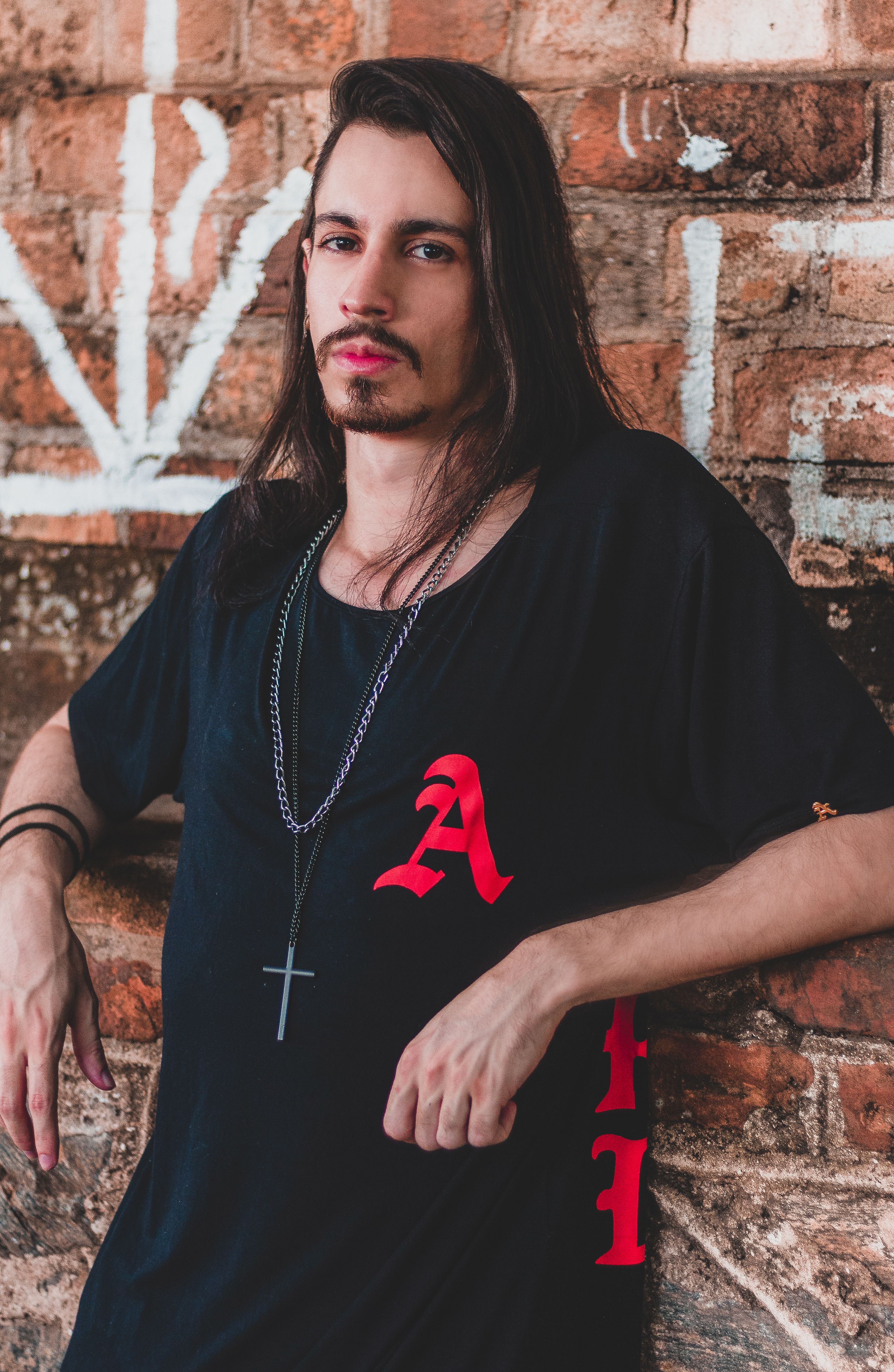 A cool, serious portrait of a man with long hair and facial hair standing against a graffiti brick wall.