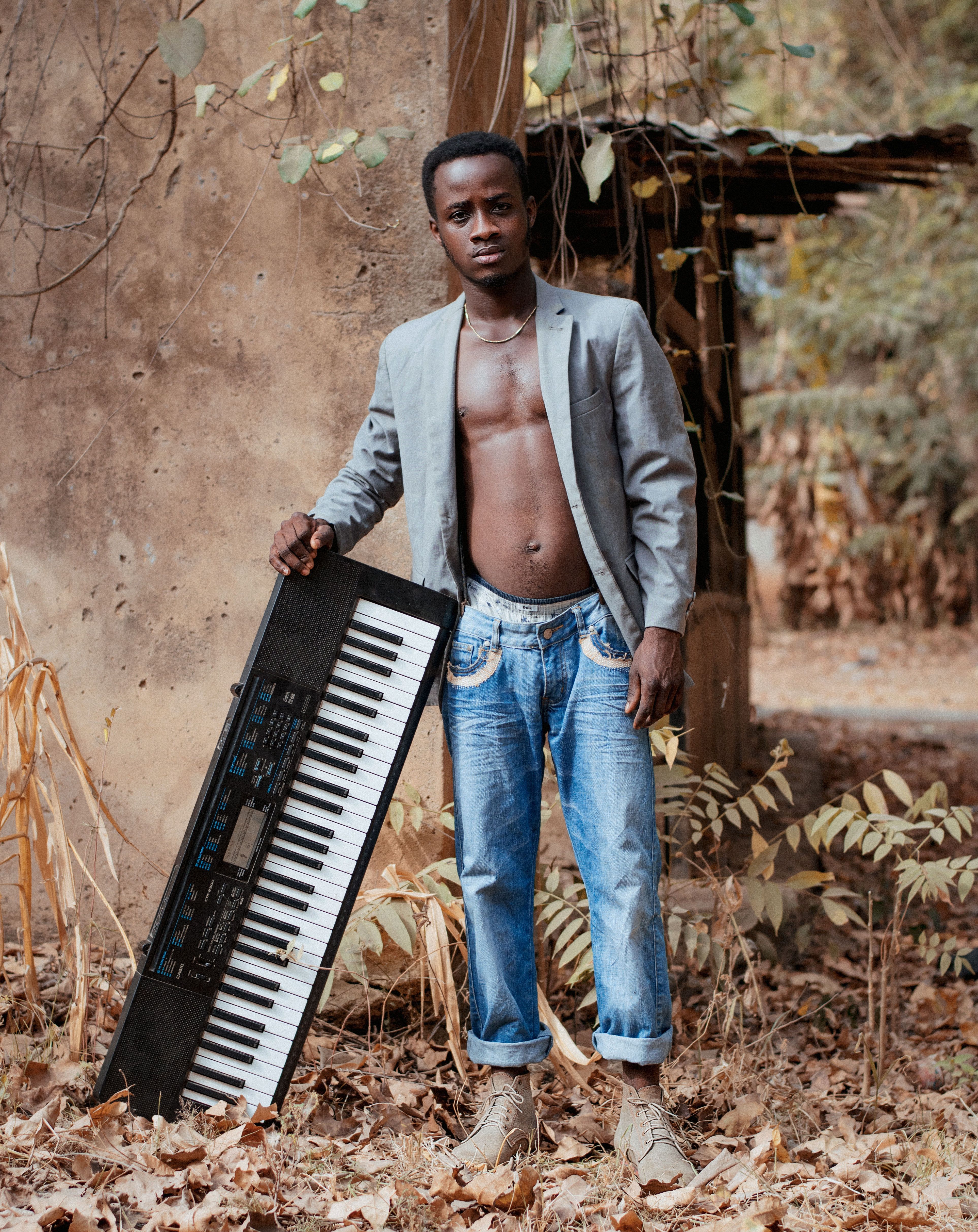 A man in a blazer and jeans poses with an electronic keyboard in a rustic outdoor setting.