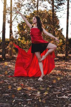 Full body of young slim barefoot female dancer in bright dress standing on one leg with raised arm and looking away while performing ballet pose on dry terrain behind trees