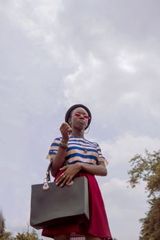 From below of fashionable young African American lady in trendy outfit and sunglasses standing in park with bag in hand against cloudy sky
