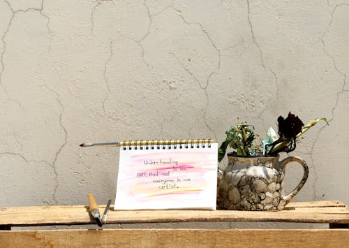 Paper notebook with written inscription near assorted paint brushes and decorative vase with plants on old wooden table near concrete shabby wall with cracks in sunlight