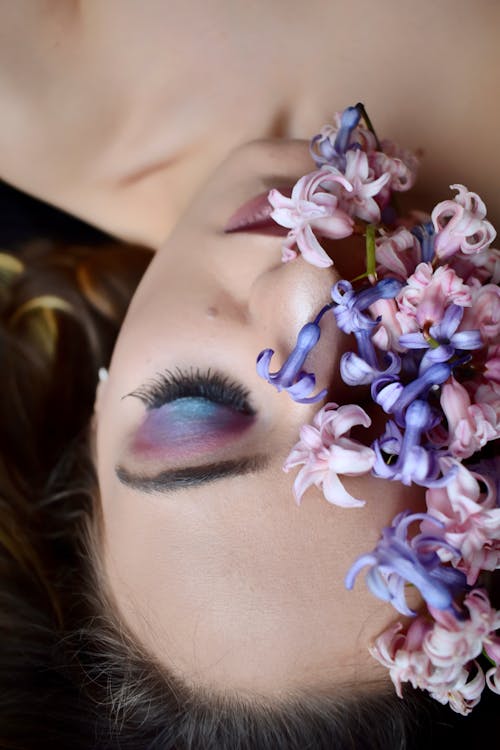 From above of female with makeup on eyelids and lips lying with bright blossoming flowers with gentle petals covering half face